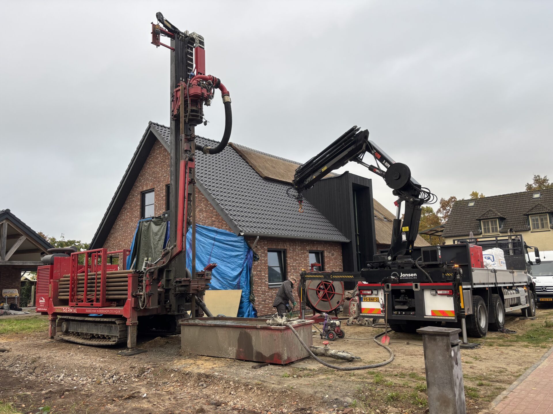 Een rode boorinstallatie en een kraanwagen voeren werkzaamheden uit naast een moderne woning voor een bodemenergiesysteem