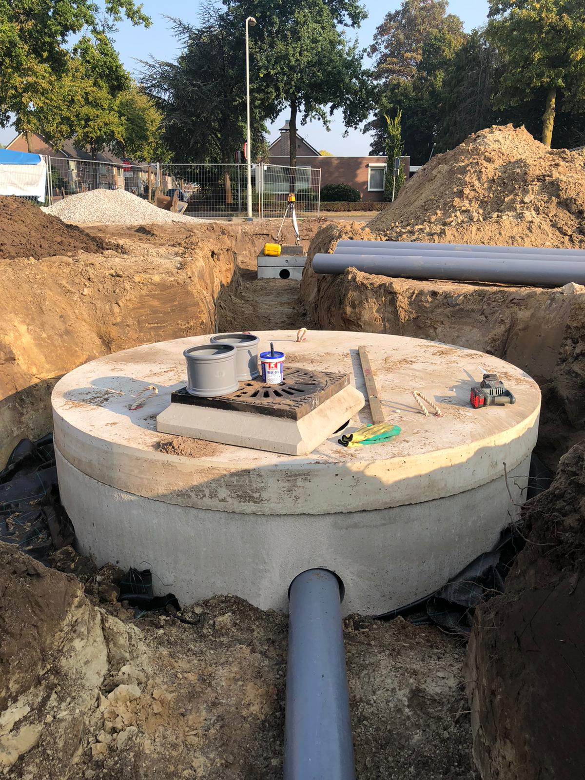 Een ronde betonnen infiltratieput met een putdeksel en aangesloten PVC-buizen ligt in een sleuf op een bouwterrein in een woonwijk.